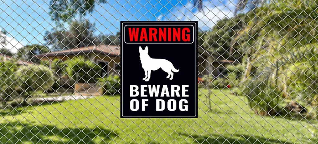 Warning sign on a chain-link fence with "Beware of Dog" and an illustration of a dog. Background shows a garden and part of a house.